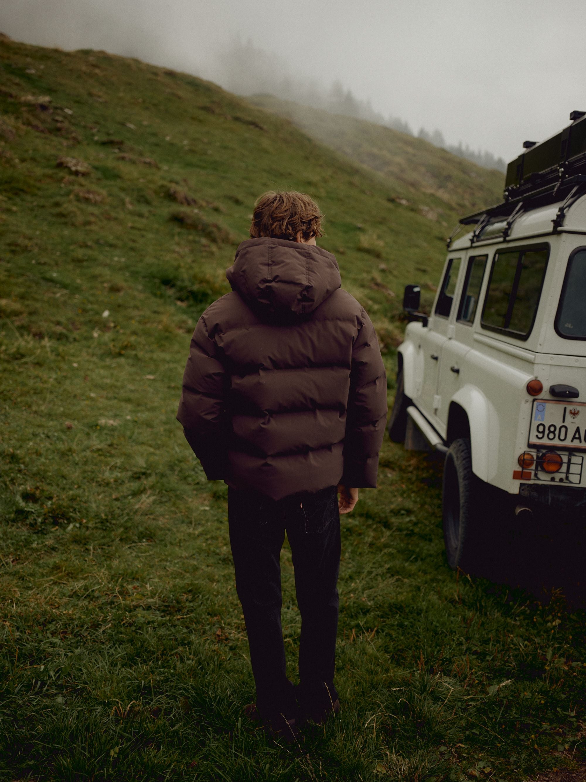 Back view of a man wearing a cocoa bean CASPARA Boreal puffer jacket outdoors next to an off-road vehicle.